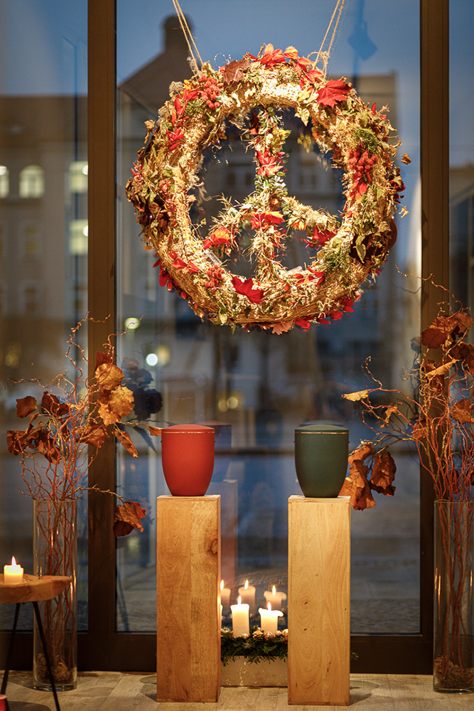 Schaufenster Bestatter Leipzig, trockene Blätter, Urnen auf Podest, Kerzen, gebundener Kranz als Friedenssymbol aus Band und Blättern